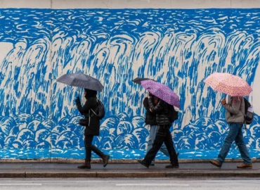 Quatre personnes marchent sous la pluie le long d'un trottoir à Porto au Portugal. Ils sont abrités par des parapluies colorés, et longent un mur bleu et blanc.